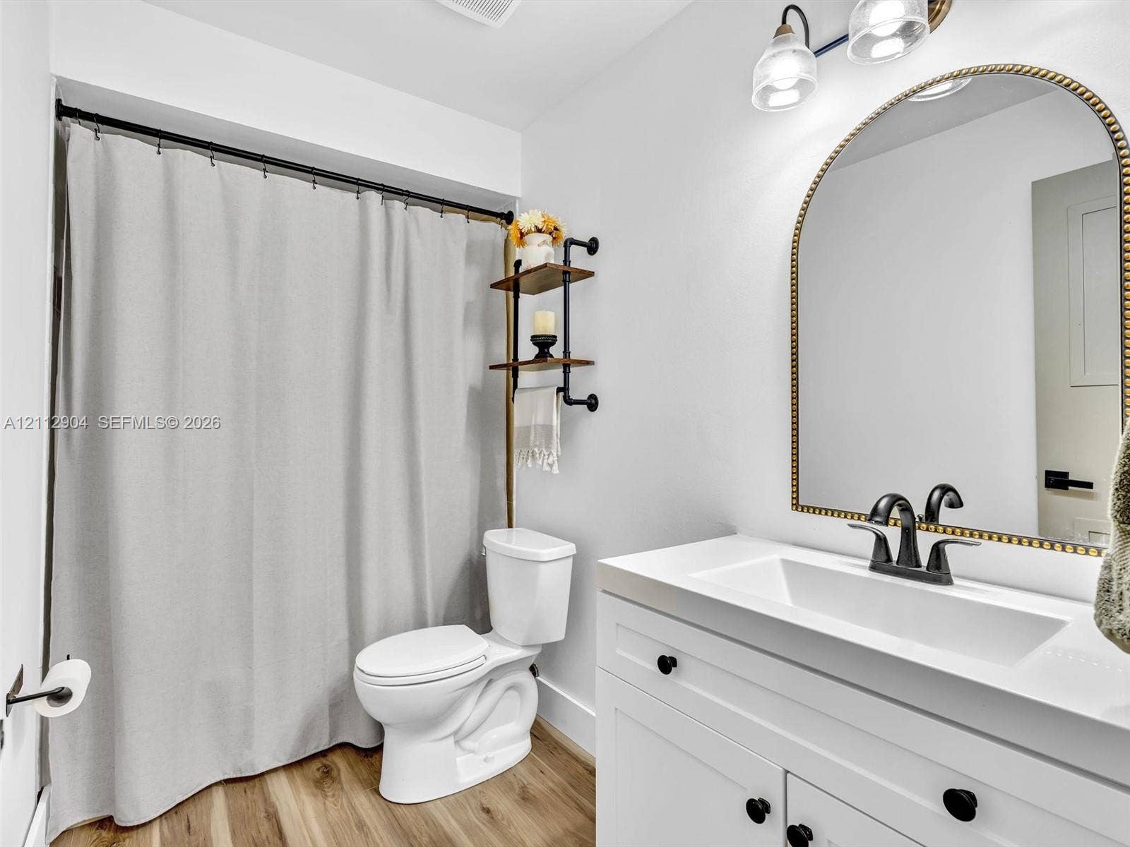 76 Forest Circle, Cooper City, FL 33026 Bathroom featuring a white vanity with an integrated rectangular sink, matte black hardware, an arched mirror with metallic trim, wood-finish flooring, and industrial-style shelving