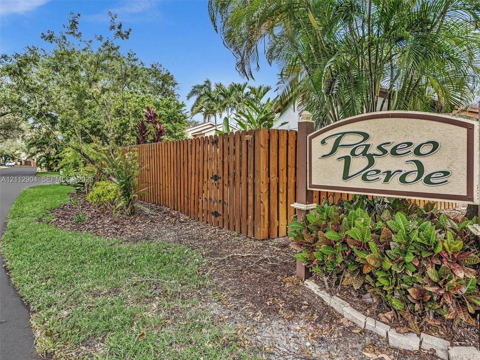 76 Forest Circle, Cooper City, FL 33026 Paseo Verde community signage featuring a custom-carved monument, vertical wood-plank fencing, asphalt-paved thoroughfare, and manicured tropical landscaping