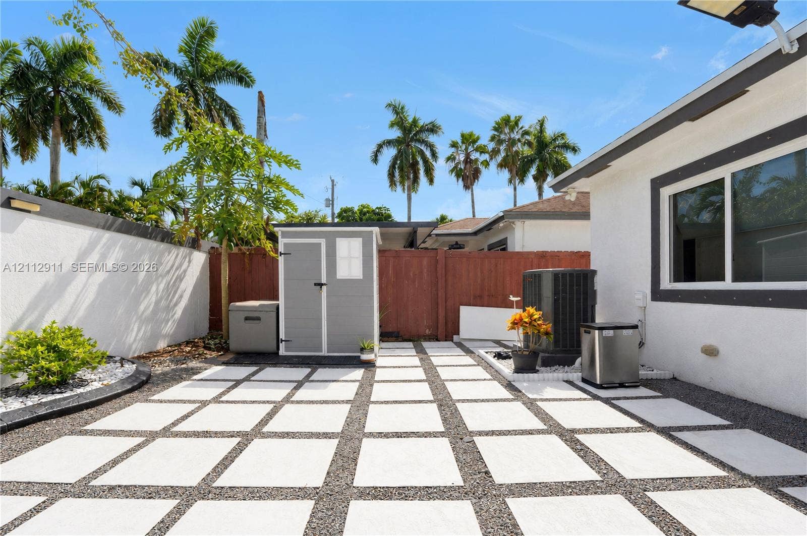 217 SW 15th Place, Homestead, FL 33030 Rear outdoor area featuring a grid-patterned concrete paver patio with gravel fill, enclosed by a white stucco perimeter wall and wood privacy fencing