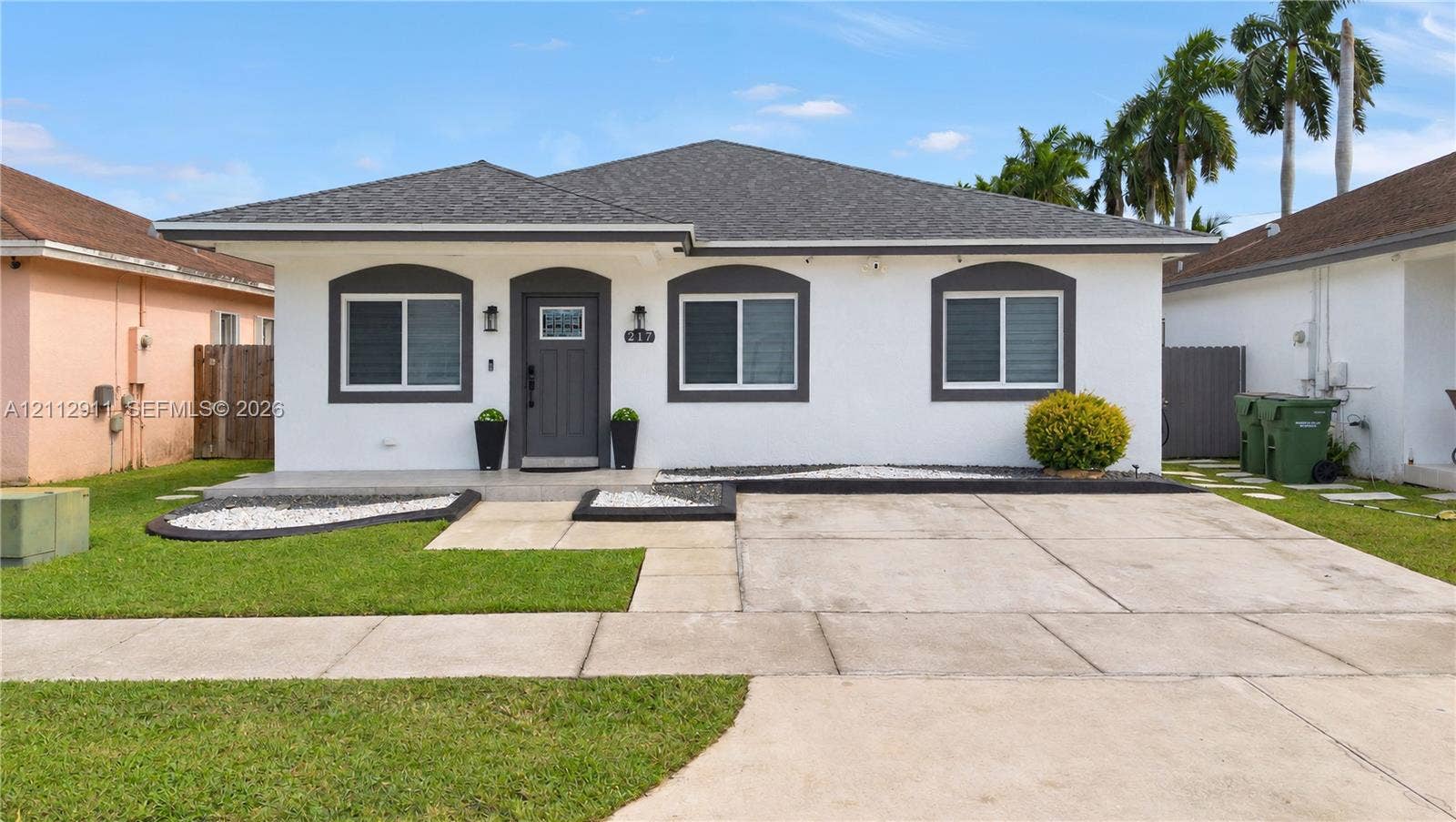 217 SW 15th Place, Homestead, FL 33030 Single-story exterior featuring a white stucco facade, contrasting dark gray trim, a concrete driveway, and landscaped front beds with stone accents