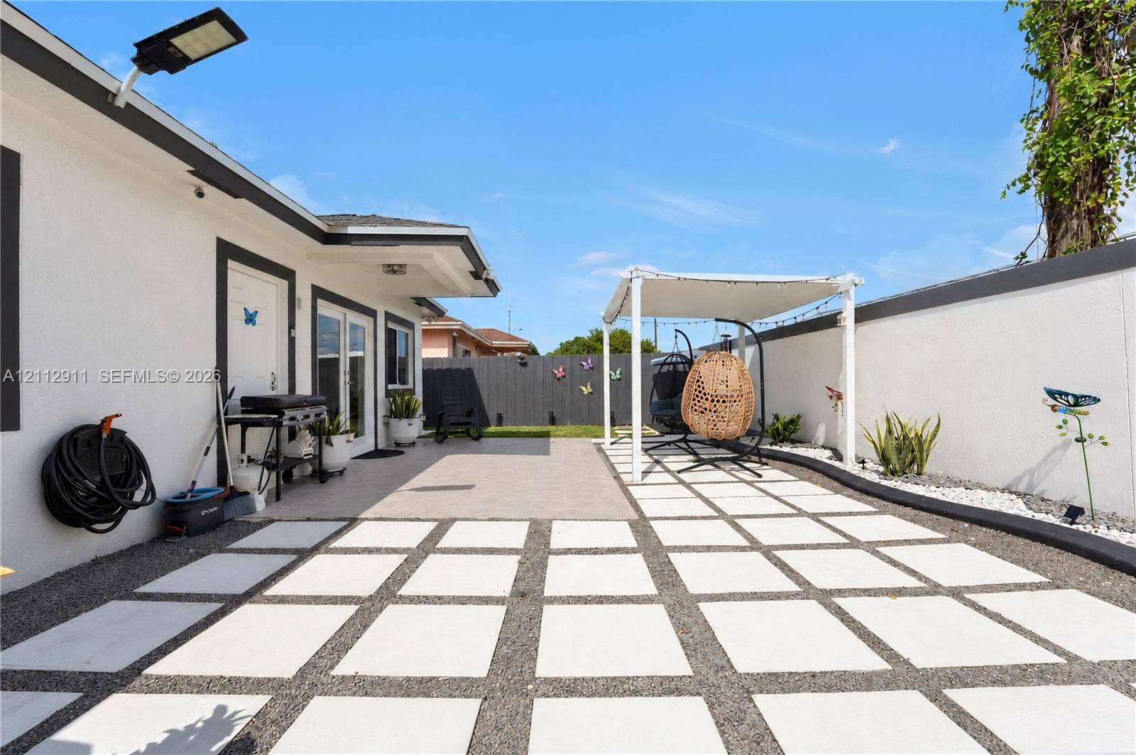 217 SW 15th Place, Homestead, FL 33030 Rear exterior featuring a large concrete paver patio with decorative aggregate inlay, a covered pergola structure, white perimeter masonry walls, and wall-mounted exterior security lighting