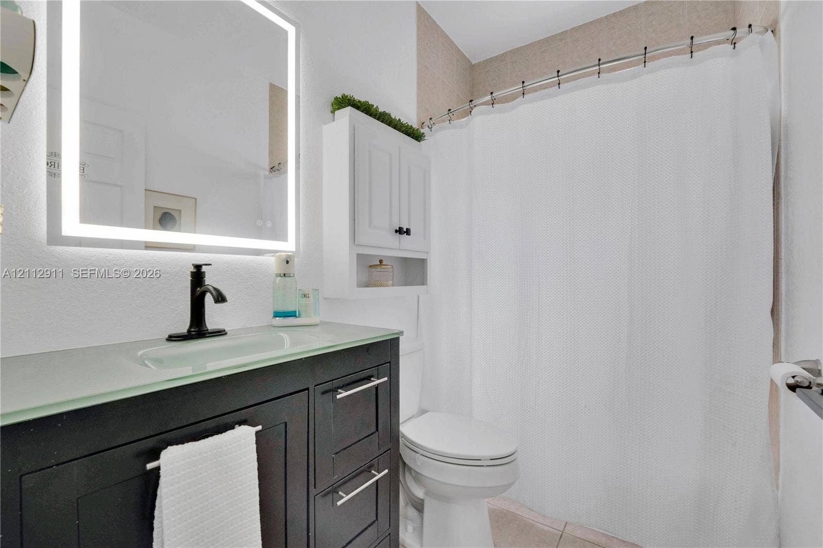 217 SW 15th Place, Homestead, FL 33030 Dark-finish vanity with a glass countertop and integrated sink, matte black faucet, wall-mounted illuminated mirror, and white built-in storage cabinet