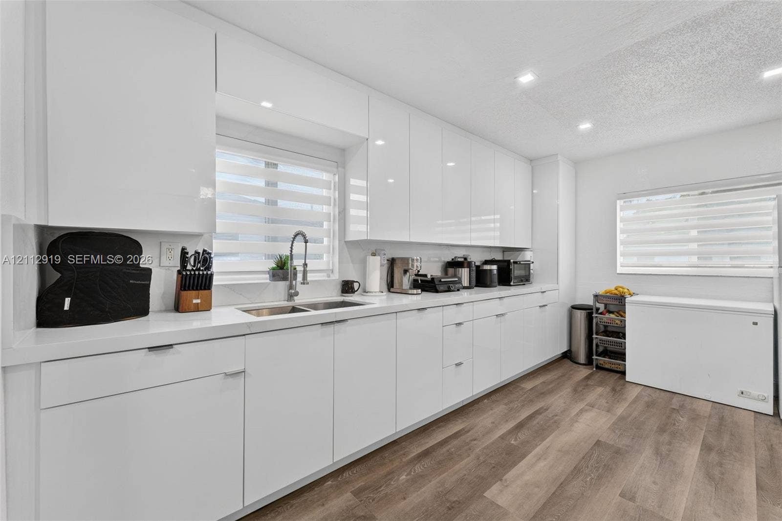 2120 NW 28th Terrace, Fort Lauderdale, FL 33311 Modern kitchen featuring high-gloss white cabinetry, white stone-surface countertops, wood-finish flooring, undermount dual-basin stainless steel sink, and recessed ceiling lighting