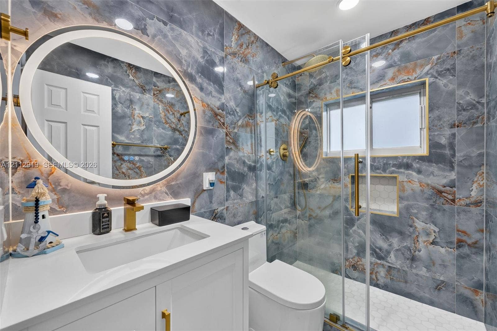 2120 NW 28th Terrace, Fort Lauderdale, FL 33311 Bathroom featuring floor-to-ceiling blue veined stone-look wall tile, backlit circular vanity mirror, white vanity with rectangular under-mount sink, brass-finish fixtures, and a glass-enclosed shower with gold-tone hardware and built-in niche