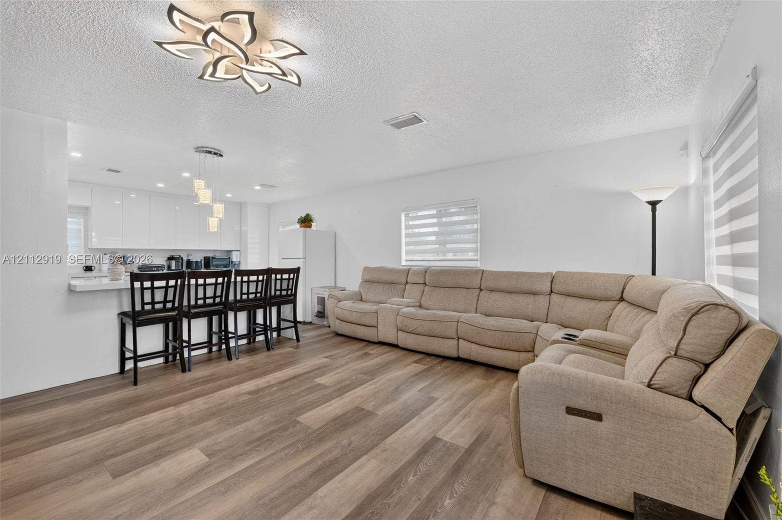 2120 NW 28th Terrace, Fort Lauderdale, FL 33311 Open-concept interior featuring wood-finish flooring, a modern geometric ceiling light fixture, high-gloss white cabinetry, and multi-layered window shades
