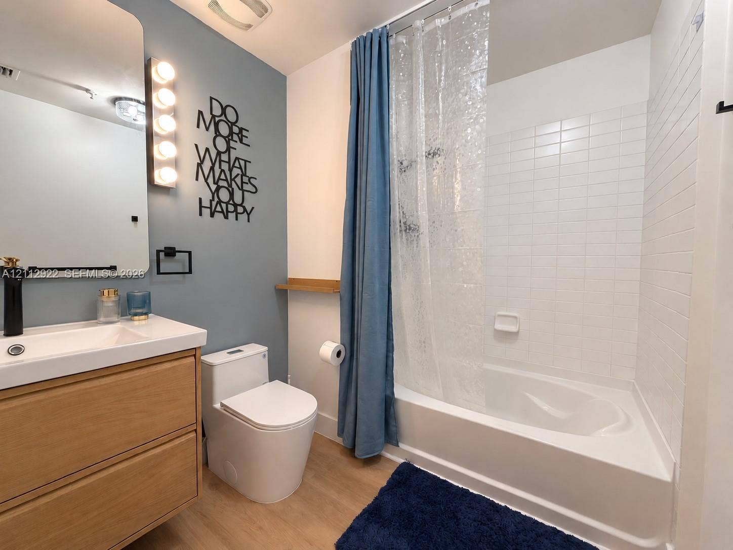 Bathroom featuring a wood-finish vanity with a rectangular sink, wall-mounted light fixture, mirror with rounded corners, and a tiled shower-tub combination