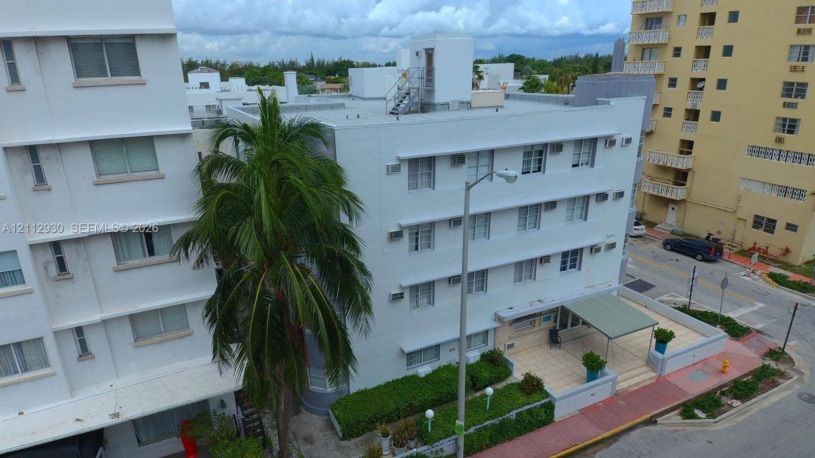 3030 Collins Avenue, #1G, Miami Beach, FL 33140 Mid-rise exterior featuring a white stucco facade, flat roofline, mature palm tree, covered entryway, and landscaped ground-level shrubbery