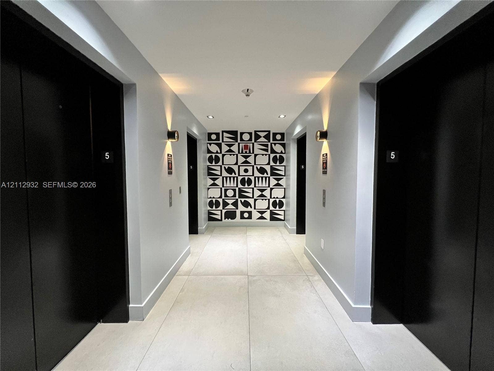 121 NE 34th Street, #903, Miami, FL 33137 Hallway featuring large-format light-toned floor tiling, matte black door finishes, geometric decorative wall paneling, and wall-mounted cylindrical sconces