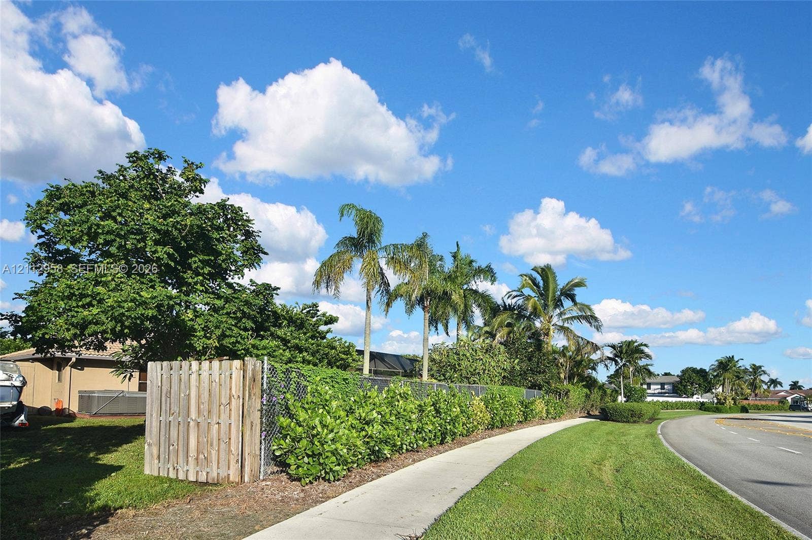 Exterior lot featuring a wooden privacy fence, chain-link perimeter, established landscaping with mature palm trees, and a concrete sidewalk