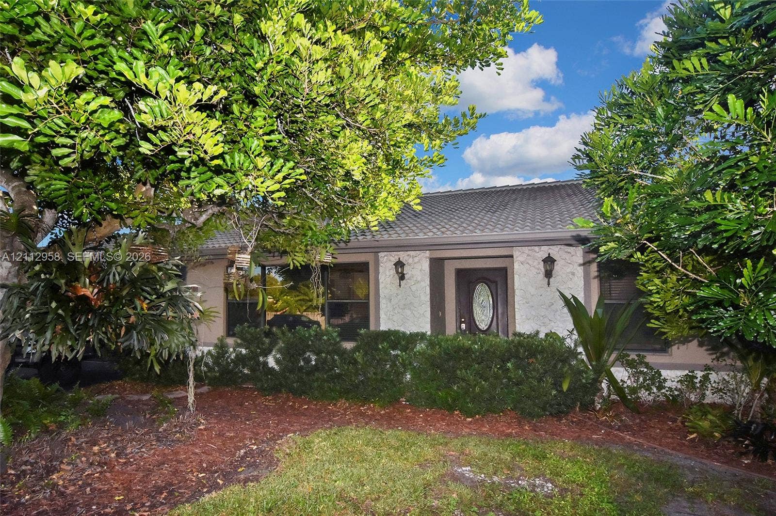 Exterior facade featuring a textured stone finish, tiled roof, decorative front door with an oval glass insert, and dual wall-mounted coach lights