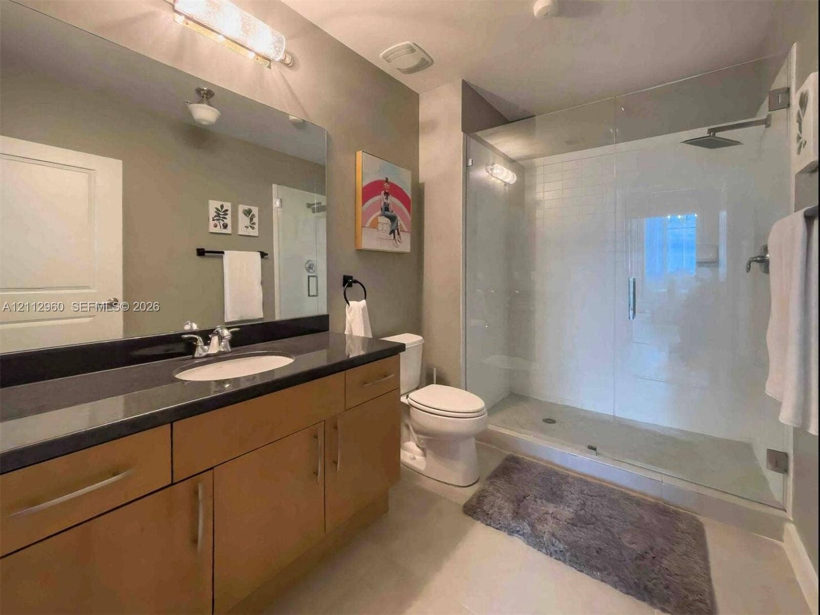 Bathroom featuring wood-finish cabinetry, dark countertop with an undermount sink, full-width mirror, and a walk-in shower with a glass enclosure and rainfall showerhead