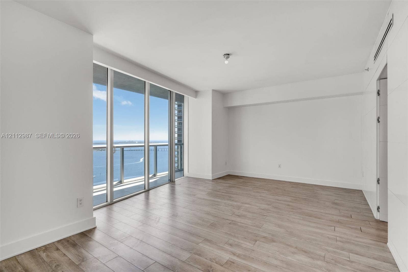 Floor-to-ceiling windows, wood-finish flooring, balcony with glass railing, and water backdrop