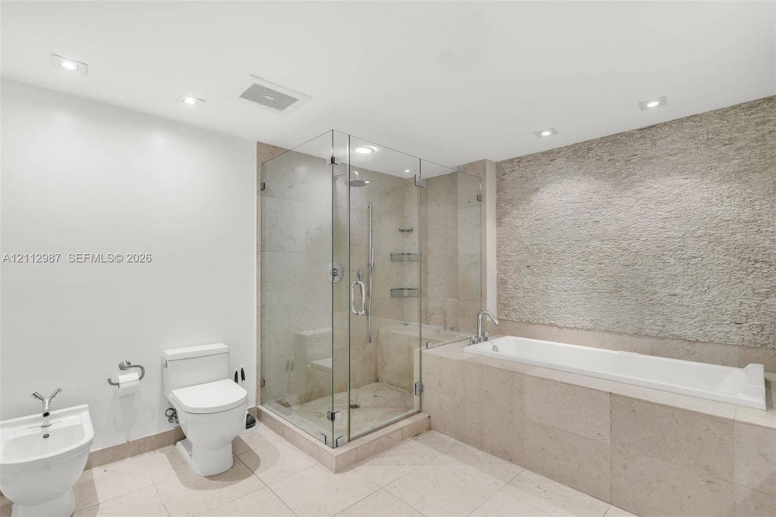 Bathroom featuring a frameless glass shower enclosure, a soaking tub with stone-tiled surround, a textured stone accent wall, recessed lighting, and a ceramic bidet and toilet