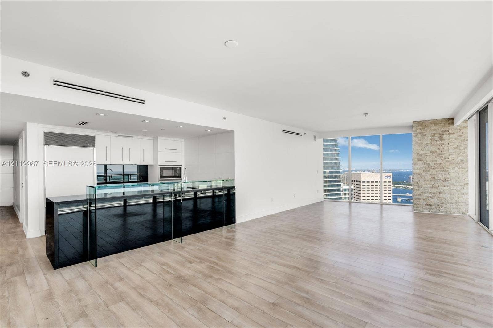 Open-concept kitchen featuring a black stone island with a glass breakfast bar, white cabinetry, integrated stainless steel appliances, wood-finish flooring, and a floor-to-ceiling brick accent wall