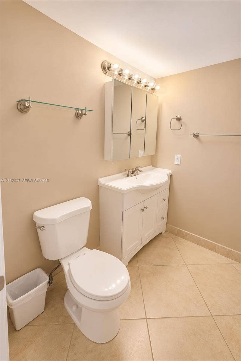 White vanity cabinet with an integrated sink, three-door mirrored medicine cabinet, multi-bulb wall-mounted lighting fixture, and neutral tile flooring