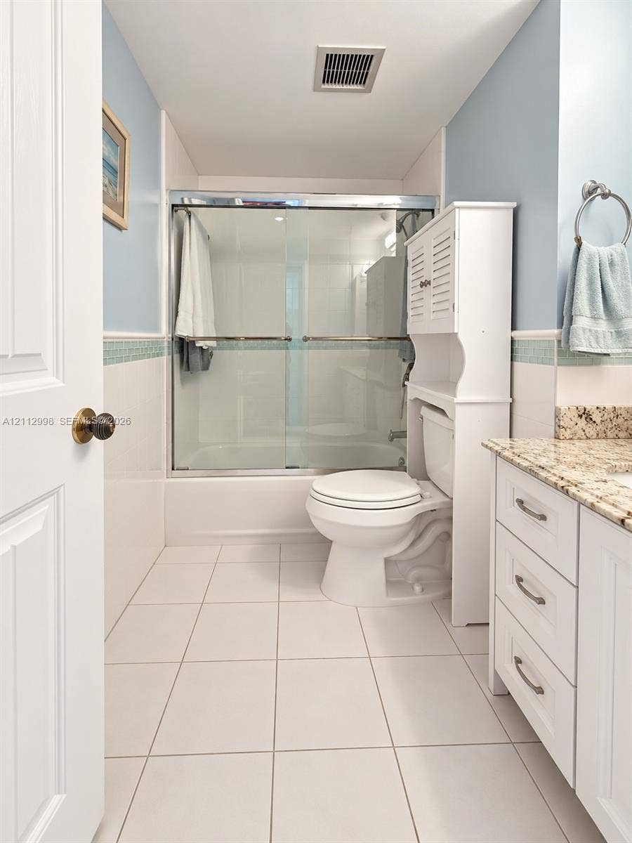 Bathroom featuring white tile flooring, a tub-shower combination with a glass sliding enclosure, a vanity with stone-finish countertop, and decorative tile trim