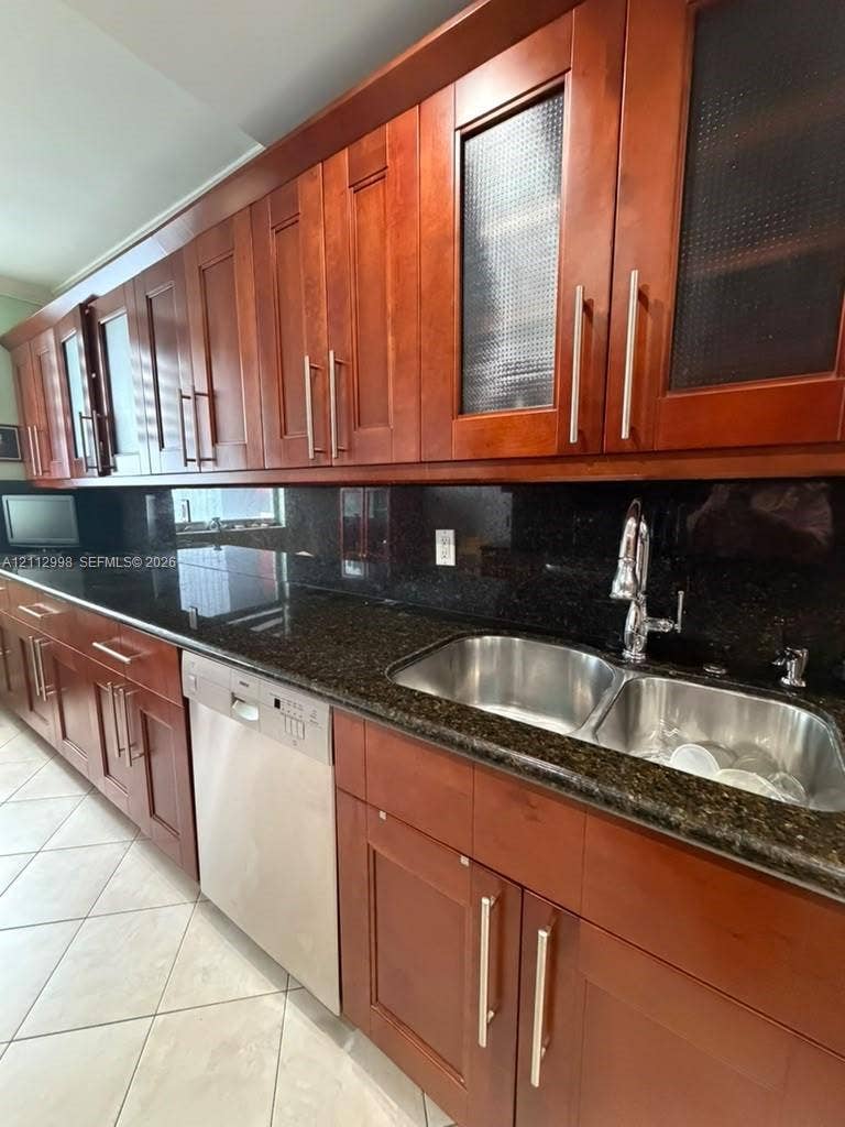 Custom wood cabinetry with glass-panel accents, dark stone countertops, stainless steel dual-basin sink with a gooseneck faucet, integrated dishwasher, and tile flooring