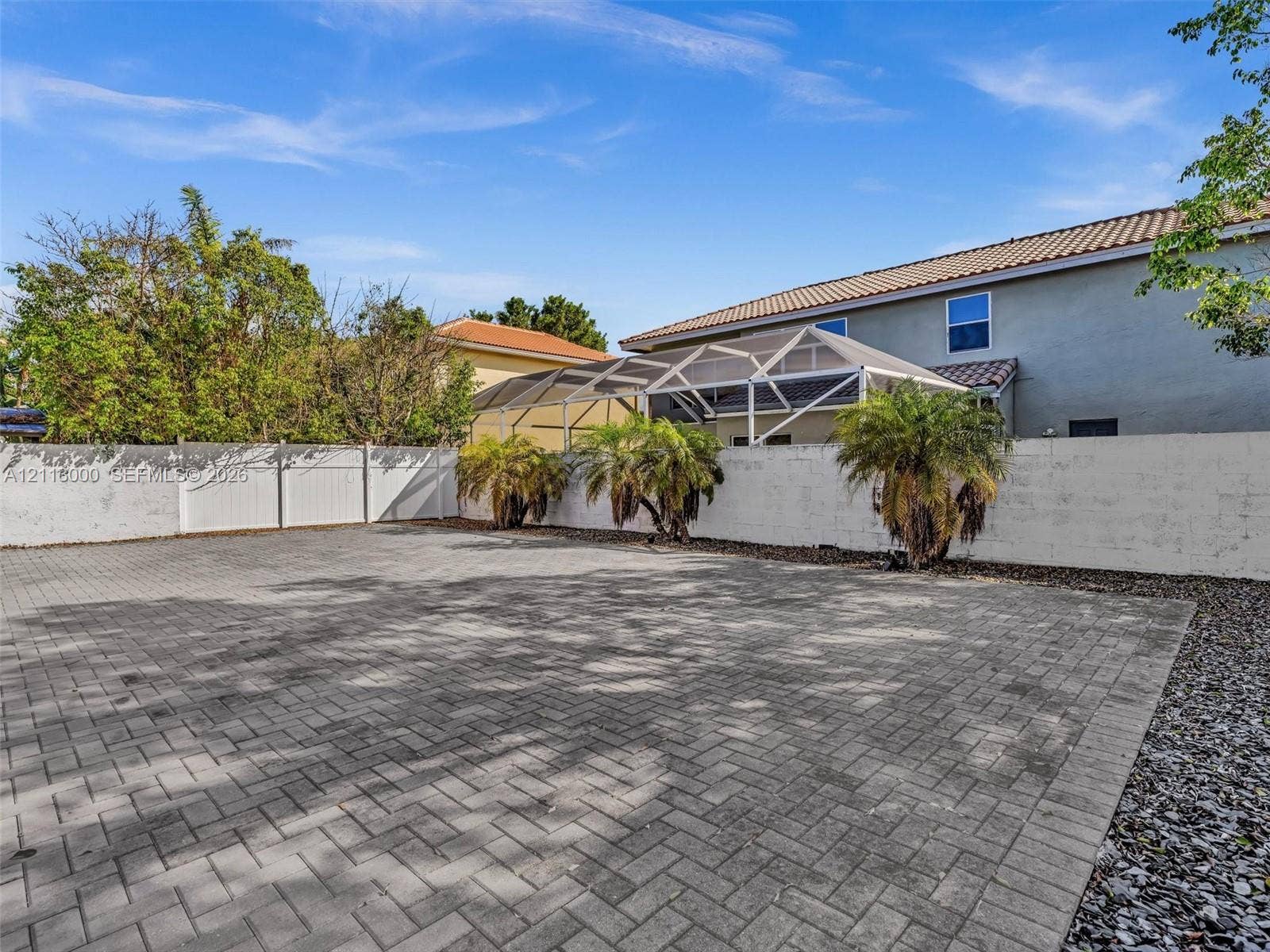 10860 La Salinas Circle, Boca Raton, FL 33428 Extensive paver patio featuring a white privacy fence, screen-enclosed patio structure, and mature palm trees