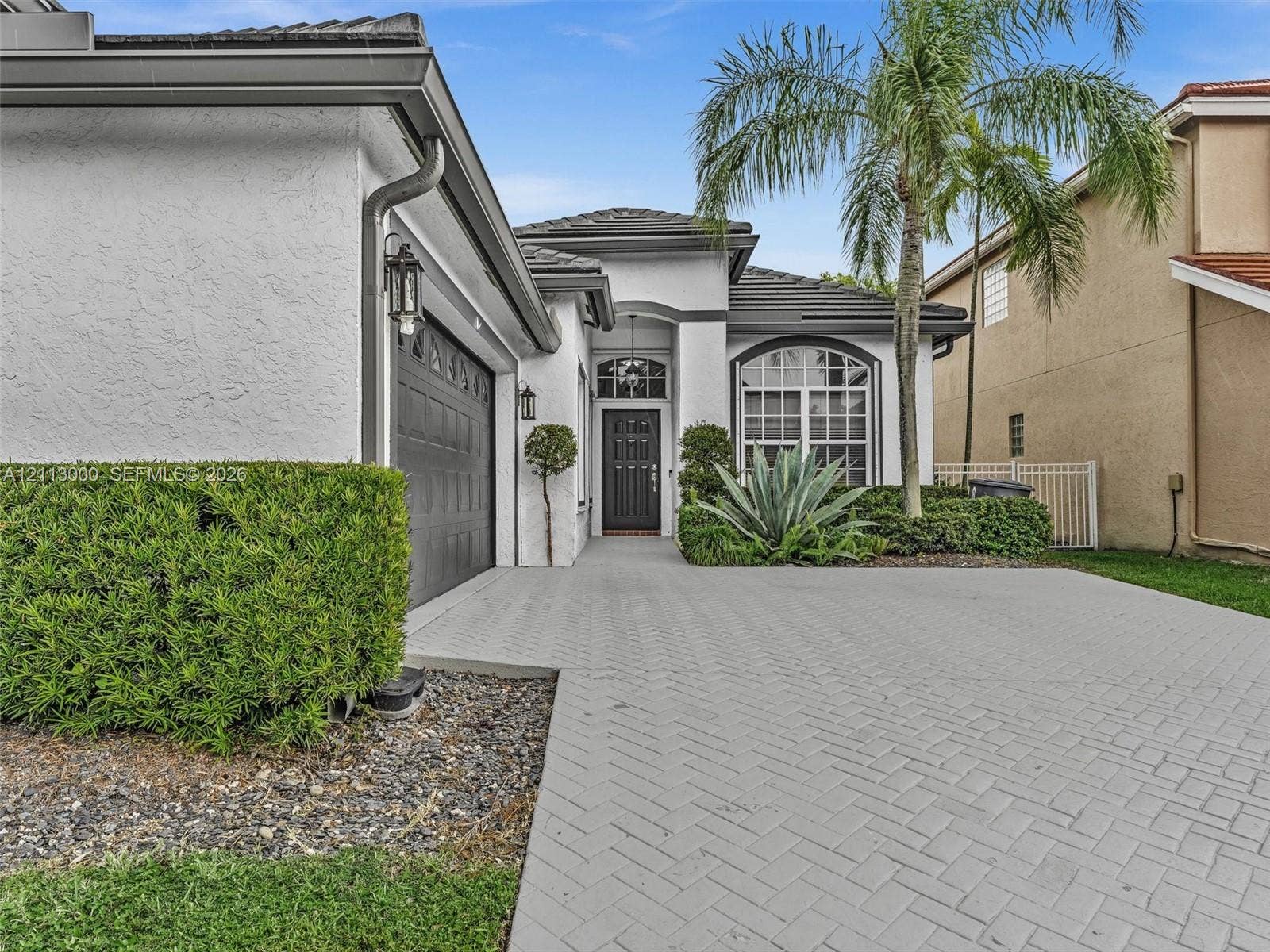 10860 La Salinas Circle, Boca Raton, FL 33428 Stucco exterior featuring a herringbone-patterned paver driveway, arched entryway, dark-tone garage door, and decorative transom window