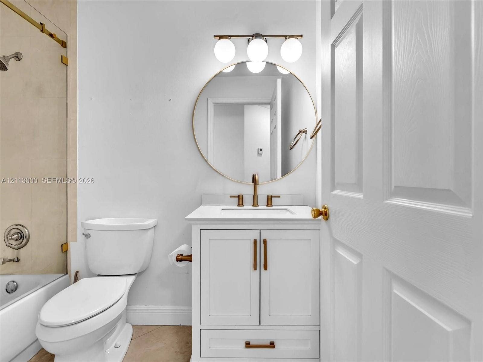 10860 La Salinas Circle, Boca Raton, FL 33428 White vanity with stone countertop, brass-toned hardware, circular mirror, three-light globe vanity fixture, and glass-enclosed shower
