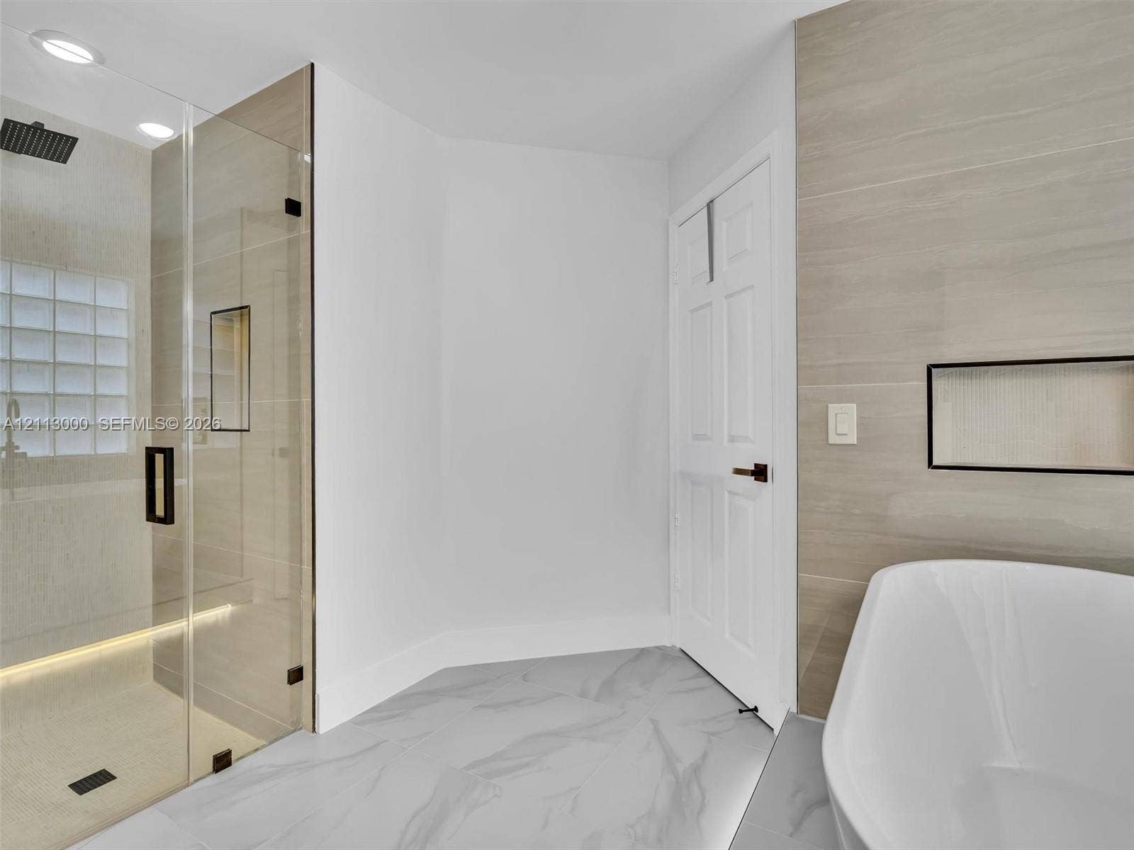 10860 La Salinas Circle, Boca Raton, FL 33428 Walk-in shower with frameless glass enclosure, rainfall showerhead, and integrated recessed shelving, featuring stone-finish flooring and a freestanding soaking tub