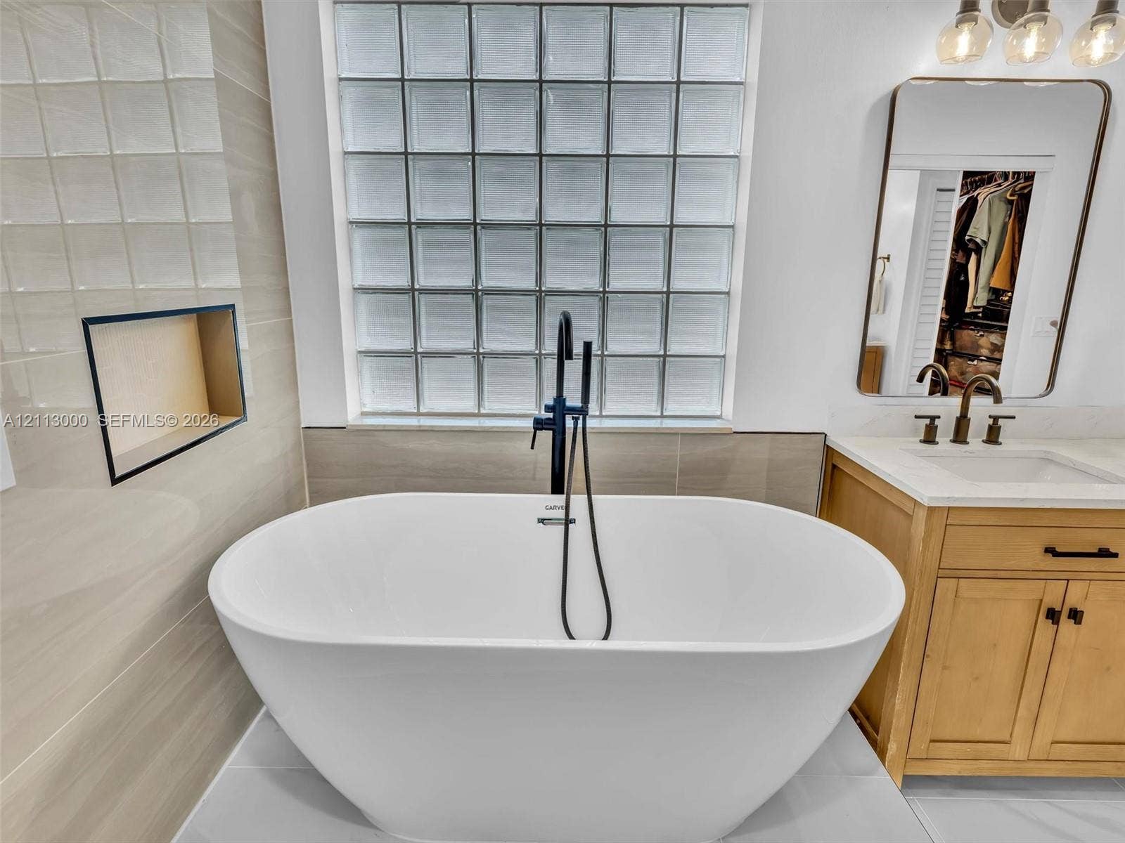 10860 La Salinas Circle, Boca Raton, FL 33428 Freestanding soaking tub featuring a matte black floor-mounted faucet with hand shower, glass block window, light-toned wood vanity with integrated countertop, and recessed tiled wall niche