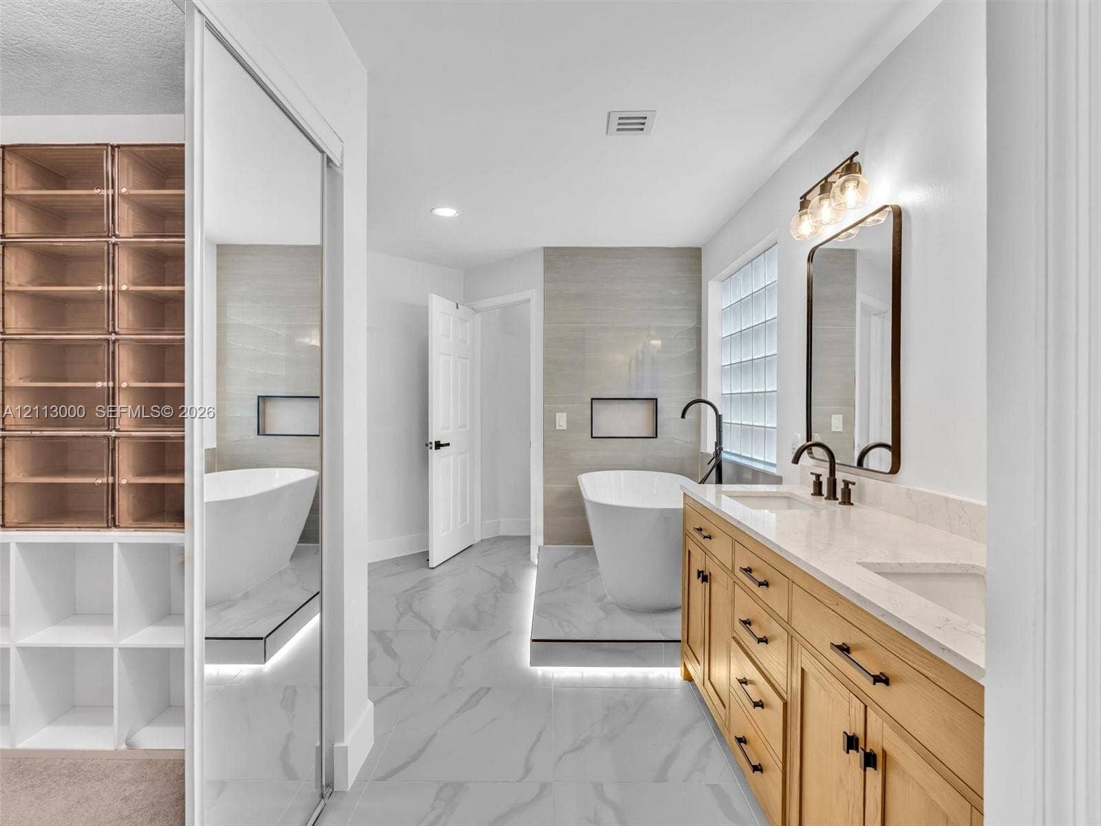10860 La Salinas Circle, Boca Raton, FL 33428 Bathroom featuring a dual-sink vanity with wood-finish cabinetry, a freestanding soaking tub set on a raised platform, built-in floor-to-ceiling shelving, large-format tile flooring, and glass block window detailing