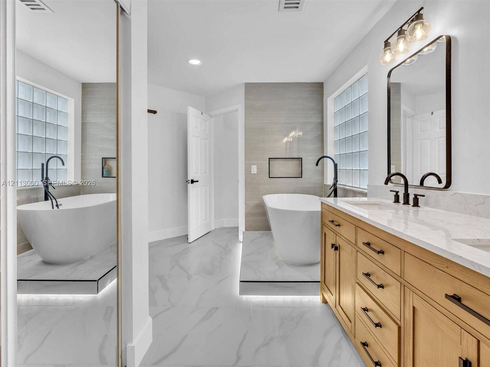 10860 La Salinas Circle, Boca Raton, FL 33428 Bathroom featuring a freestanding soaking tub on a raised platform with under-cabinet lighting, wood-finish cabinetry, quartz countertop with dual integrated sinks, and glass block windows