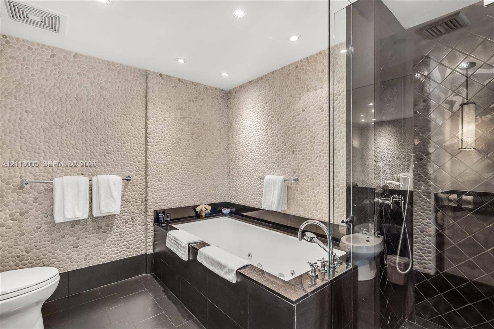 Bathroom featuring pebble-textured stone accent walls, a jetted soaking tub with a dark stone surround, a glass-enclosed shower with diamond-patterned tile, and recessed ceiling lighting