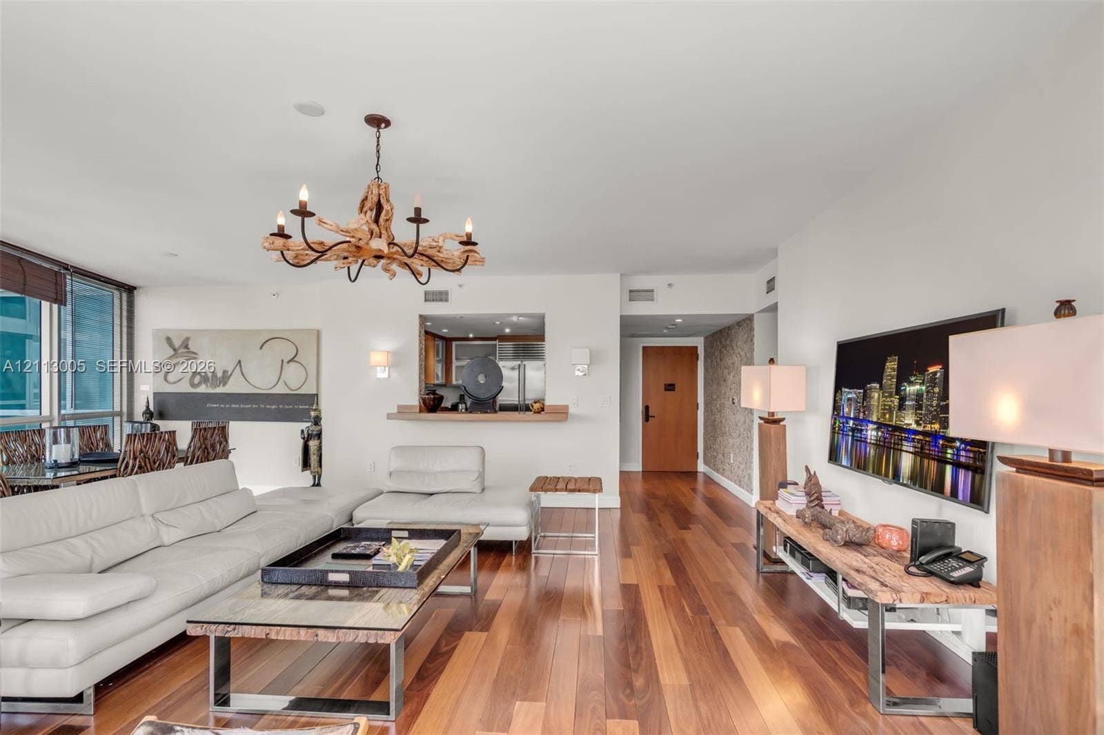 Open-concept living area featuring wood-finish flooring, a natural driftwood chandelier, textured wall accents, and a service pass-through window with a wood ledge