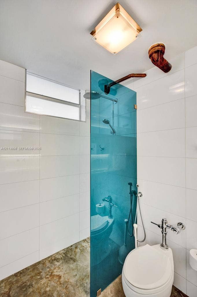 Walk-in shower enclosure with a vibrant blue glass partition, large-format white wall tiling, stone-finish flooring, rainfall shower head, and a ceiling-mounted square light fixture