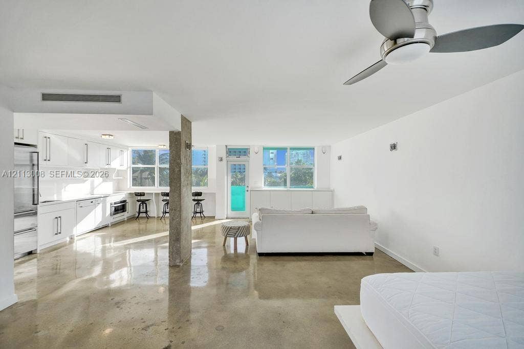 Polished concrete flooring, white cabinetry with integrated appliances, exposed concrete structural column, and a modern multi-blade ceiling fan