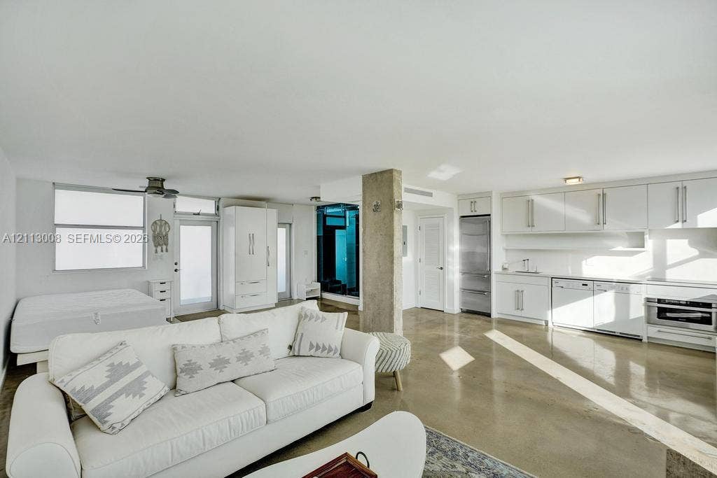 Polished concrete flooring, exposed concrete structural pillar, white cabinetry with integrated appliances, and ceiling-mounted fan