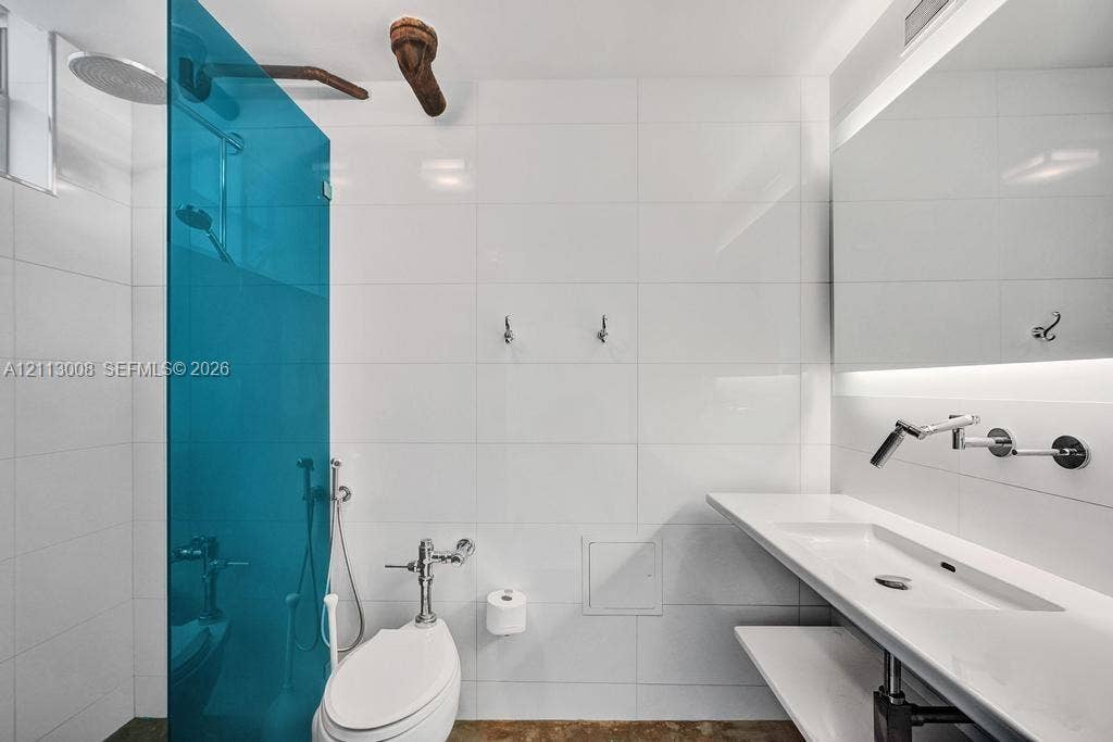 Bathroom featuring large-format white tile walls, a blue glass shower partition, wall-mounted faucet, floating countertop with integrated sink, and illuminated mirror