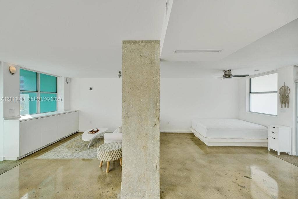 Open-concept interior featuring polished concrete flooring, a central exposed concrete structural column, high-gloss white built-in cabinetry, and a ceiling-mounted fan