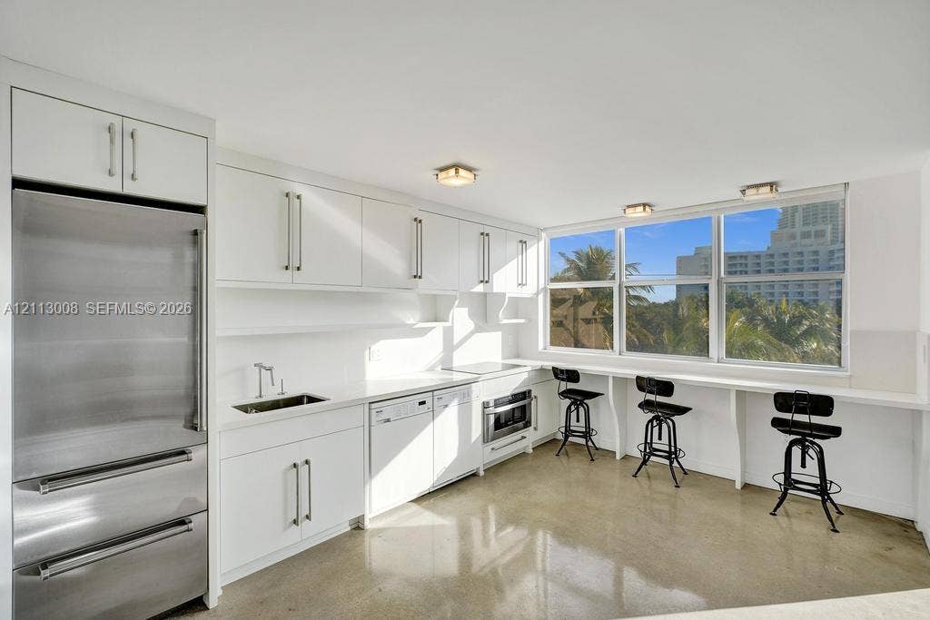 Kitchen featuring white cabinetry with integrated hardware, polished concrete flooring, stainless steel refrigerator, built-in dishwasher, and a window-lined counter