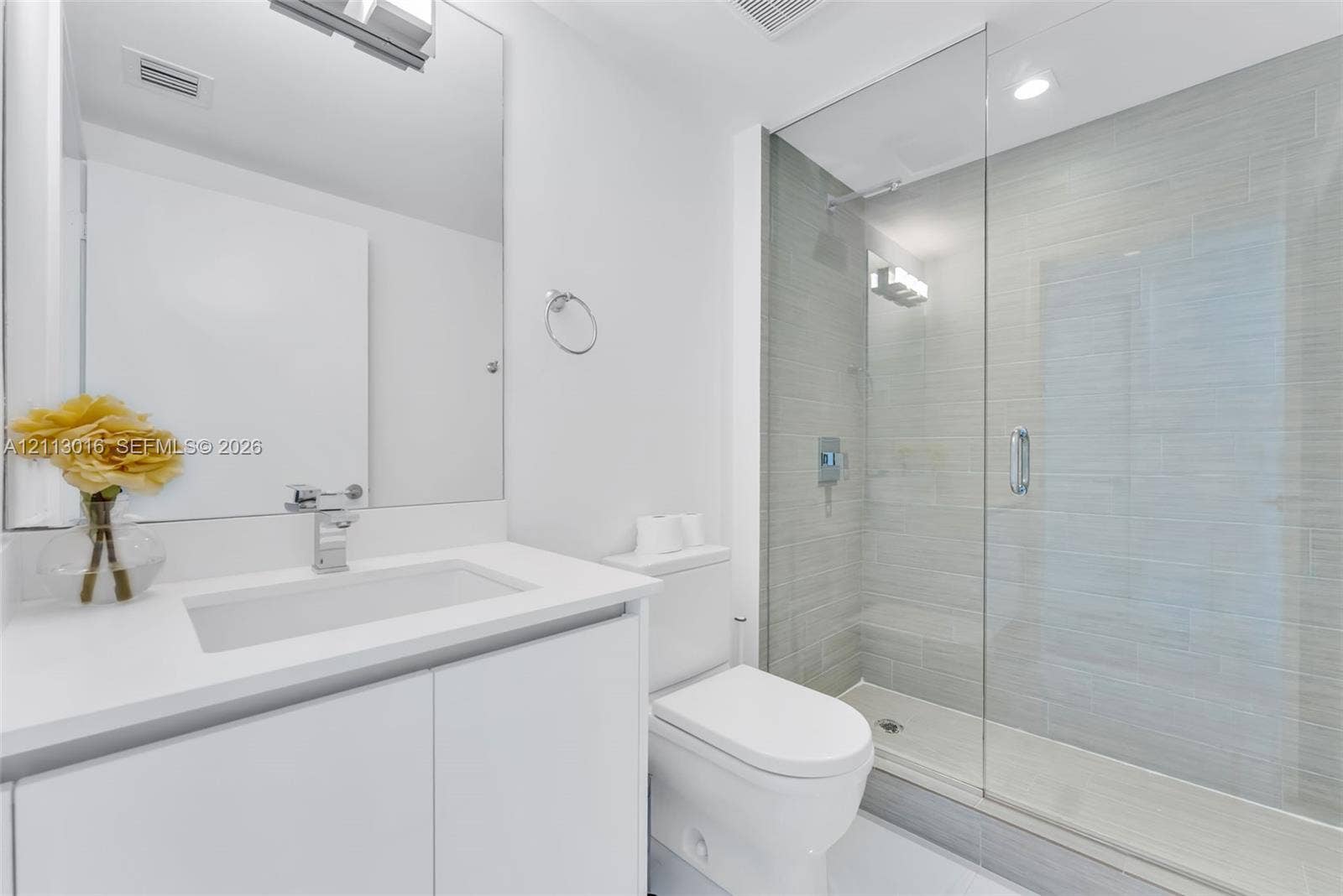 White vanity cabinet with a solid surface countertop and integrated rectangular sink, frameless glass walk-in shower with horizontal gray tile surround, and wall-mounted vanity mirror