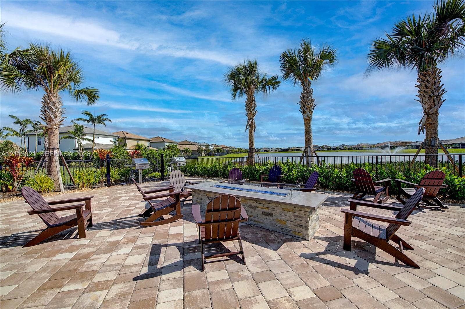 8228 Tailshot Court, Lake Worth, Lake Worth, FL 33467 Paver patio featuring a stone-clad linear fire pit, metal outdoor grill, water-facing fence line, and mature palm trees
