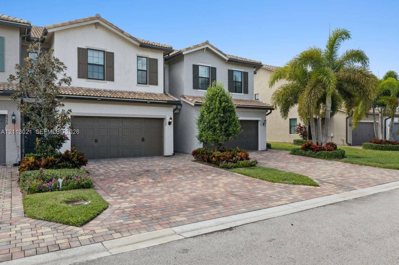 8228 Tailshot Court, Lake Worth, Lake Worth, FL 33467 Two-story multi-family exterior featuring a tiled roof, paver driveway, attached multi-car garage, and established landscaping with palm trees