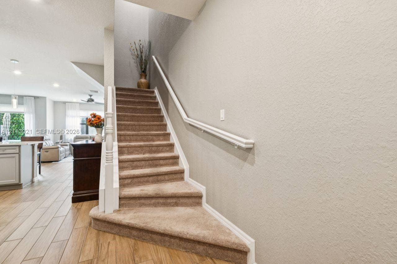 8228 Tailshot Court, Lake Worth, Lake Worth, FL 33467 Staircase featuring carpeted treads, a white wood baluster, and a wall-mounted handrail, set against wood-finish flooring