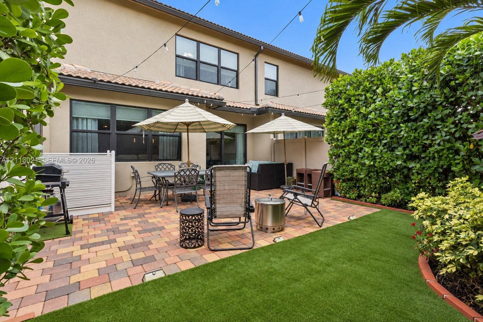 8228 Tailshot Court, Lake Worth, Lake Worth, FL 33467 Rear patio featuring multicolored paver hardscaping, synthetic turf lawn, privacy hedging, and overhead string lighting