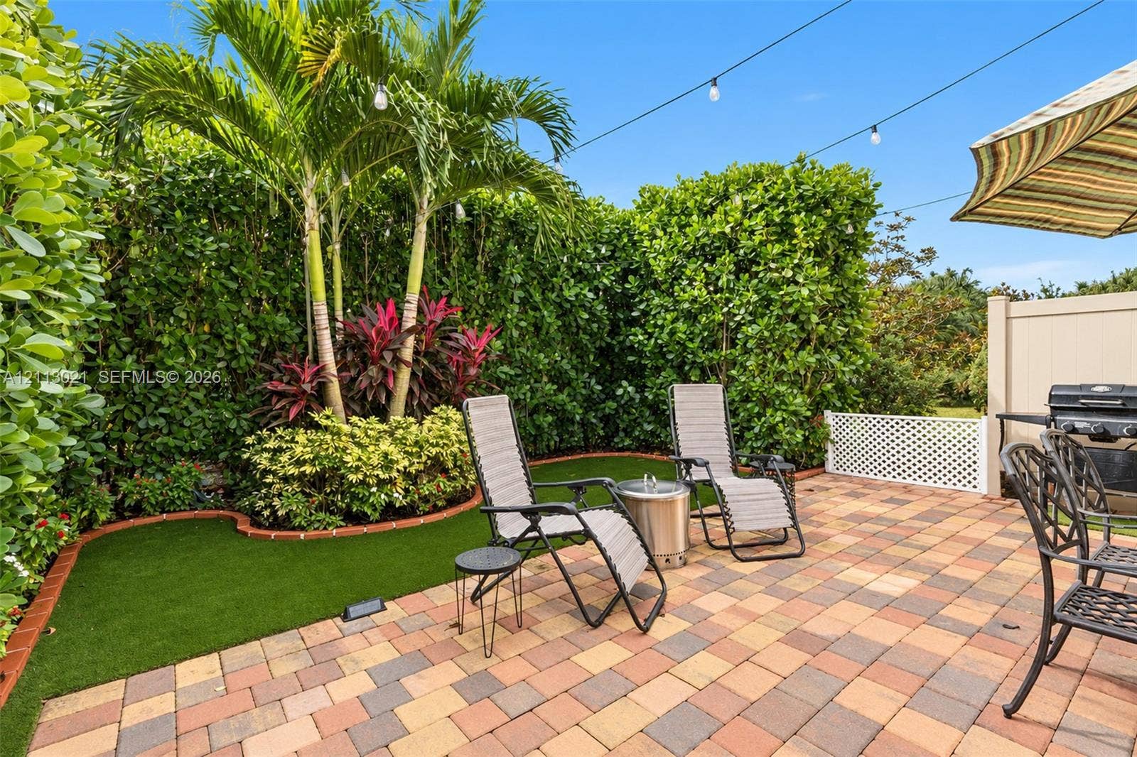 8228 Tailshot Court, Lake Worth, Lake Worth, FL 33467 Backyard featuring multicolor paver patio, synthetic turf, mature palm trees, perimeter privacy hedging, and overhead string lighting