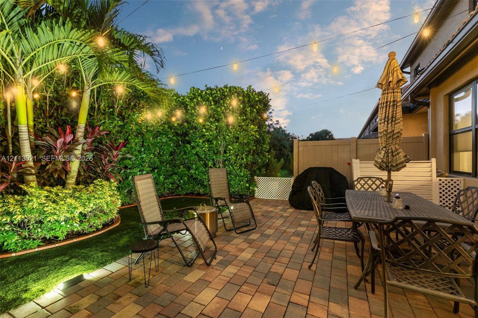 8228 Tailshot Court, Lake Worth, Lake Worth, FL 33467 Rear patio featuring multi-tone pavers, mature tropical landscaping, artificial turf, perimeter fencing, and overhead string lighting