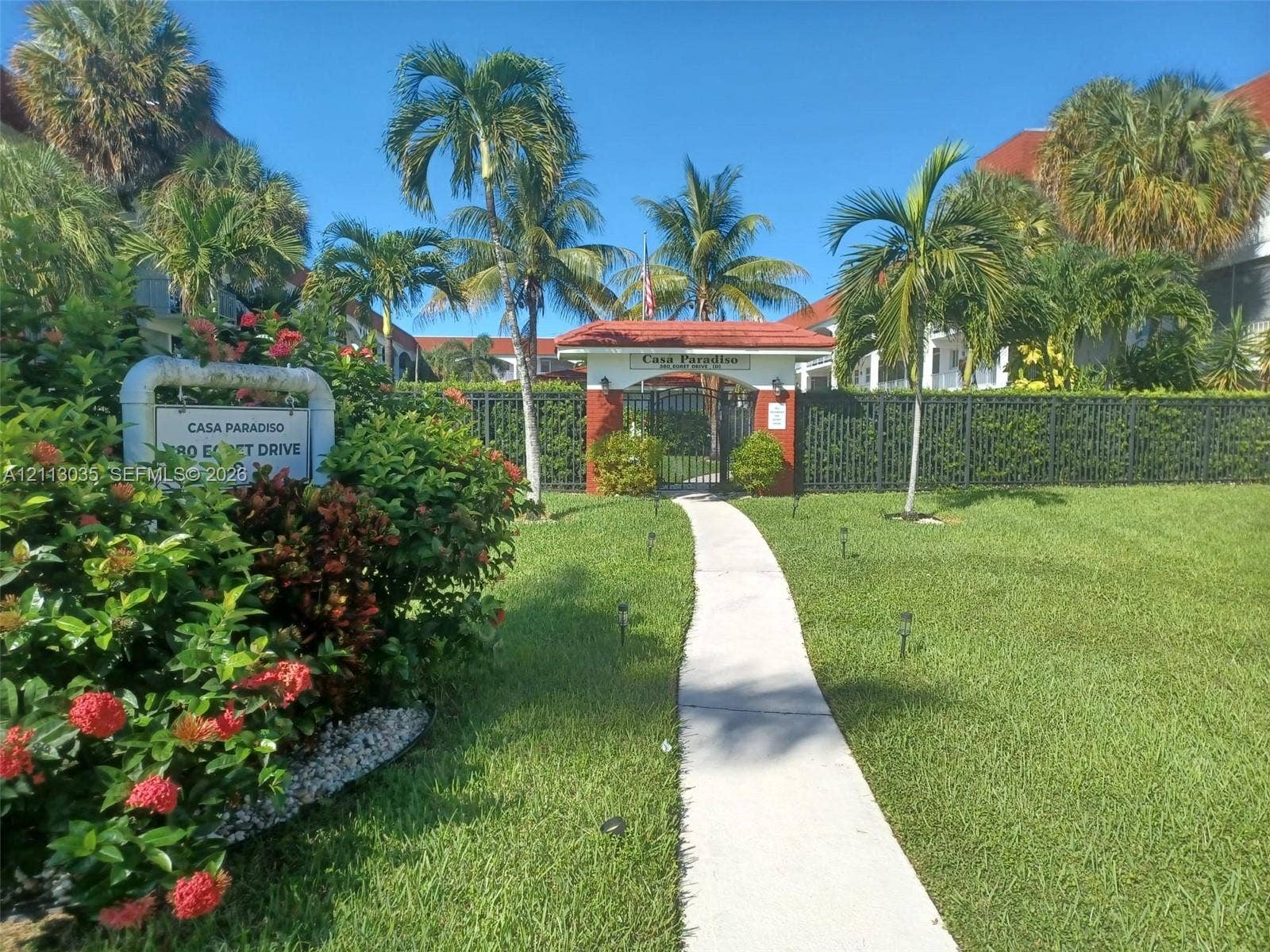 Gated community entrance featuring red-tiled roof pillars, a concrete walkway, mature palm trees, and manicured landscaping