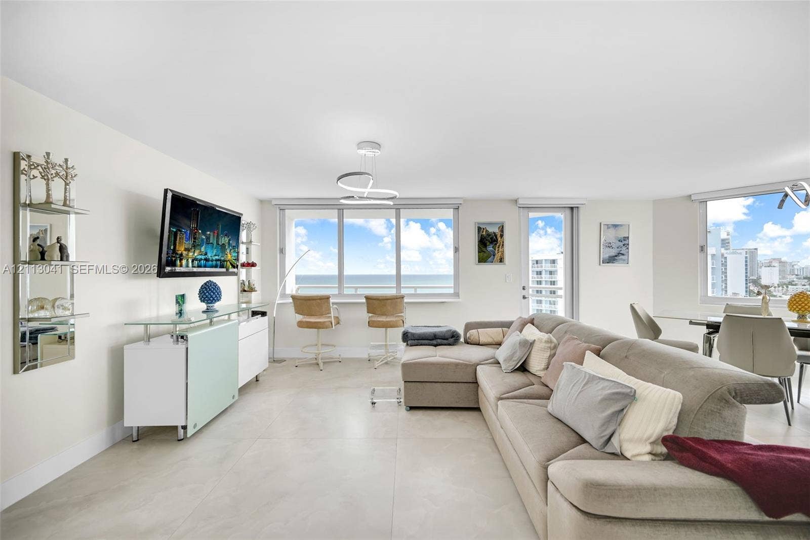 7135 Collins Avenue, #1426, Miami Beach, FL 33141 Open-concept living area featuring large-format tile flooring, a modern sculptural chandelier, floor-to-ceiling windows with coastal scenery, and a glass-paneled entry door