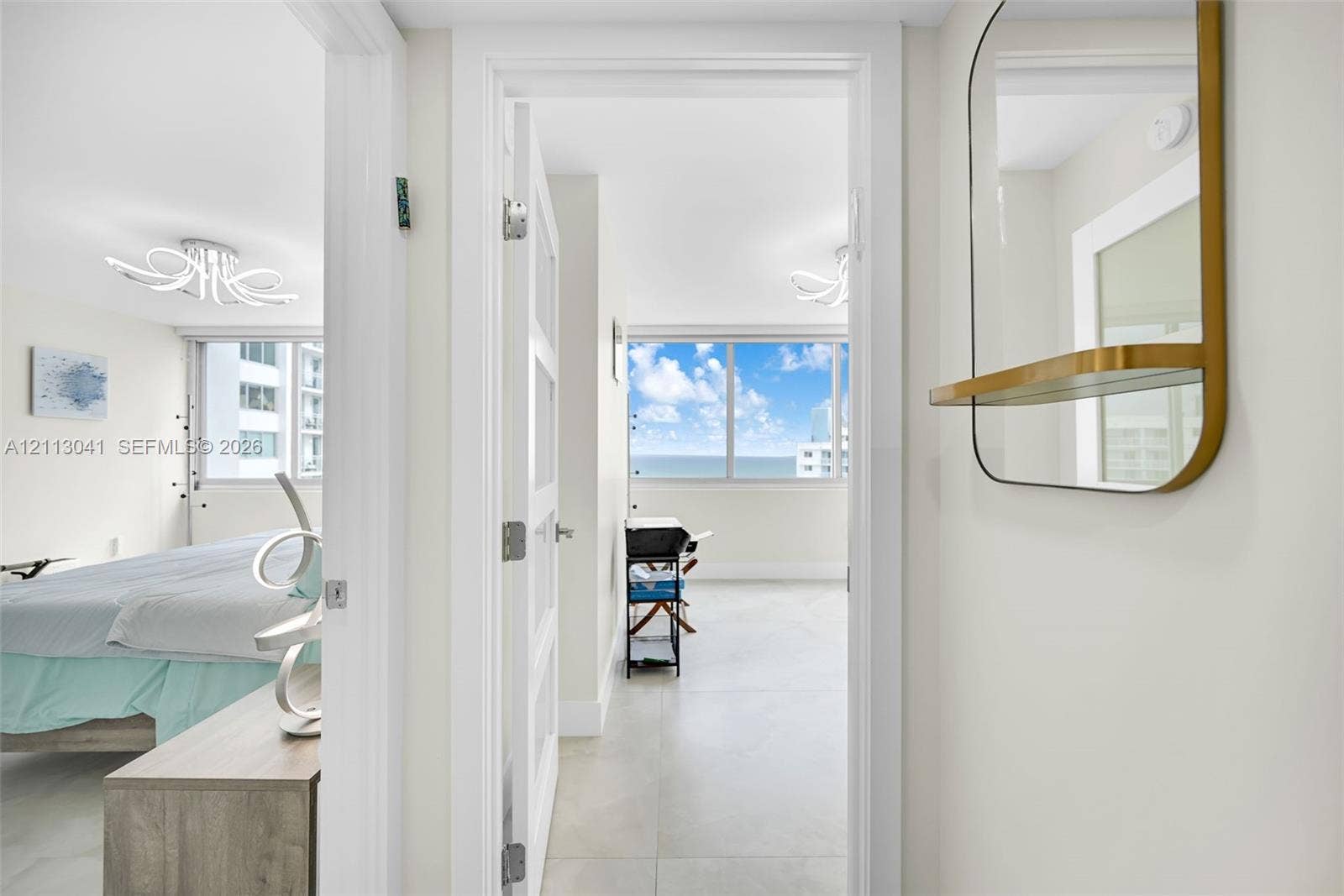 7135 Collins Avenue, #1426, Miami Beach, FL 33141 Light-toned large-format tile flooring, arched decorative ceiling lighting fixtures, a gold-framed wall-mounted mirror with an integrated shelf, and floor-to-ceiling windows with water scenery
