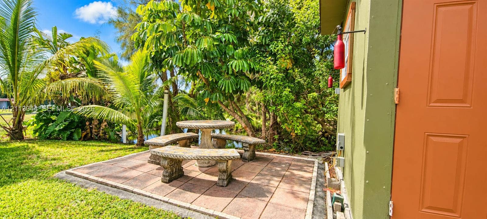 6735 NW 62nd Street, Tamarac, FL 33321 Outdoor stone paver patio featuring an integrated stone table and bench set, surrounded by established landscaping and palm trees