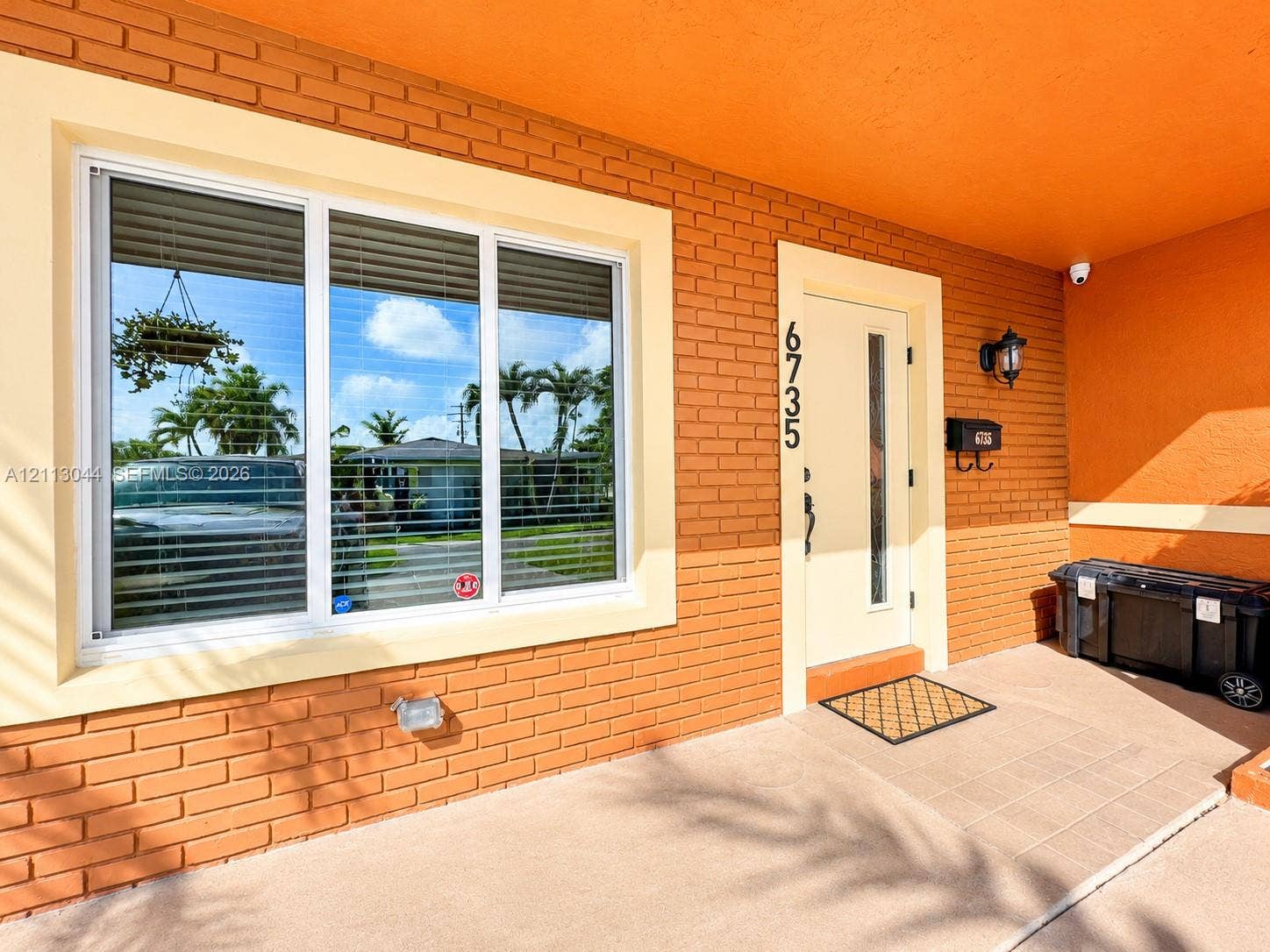 6735 NW 62nd Street, Tamarac, FL 33321 Exterior entrance featuring painted brick masonry, large picture window with wide-frame casing, solid-core door with glass inset, and integrated wall-mounted light fixture