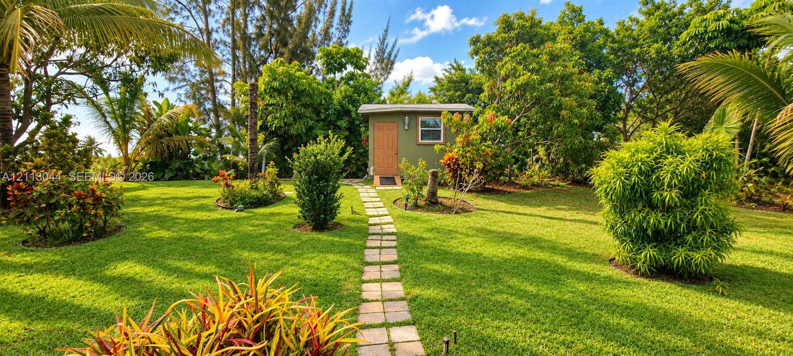 6735 NW 62nd Street, Tamarac, FL 33321 Expansive lawn featuring mature tropical landscaping, a stone-paver walkway, and a detached exterior structure with a central entry door and window
