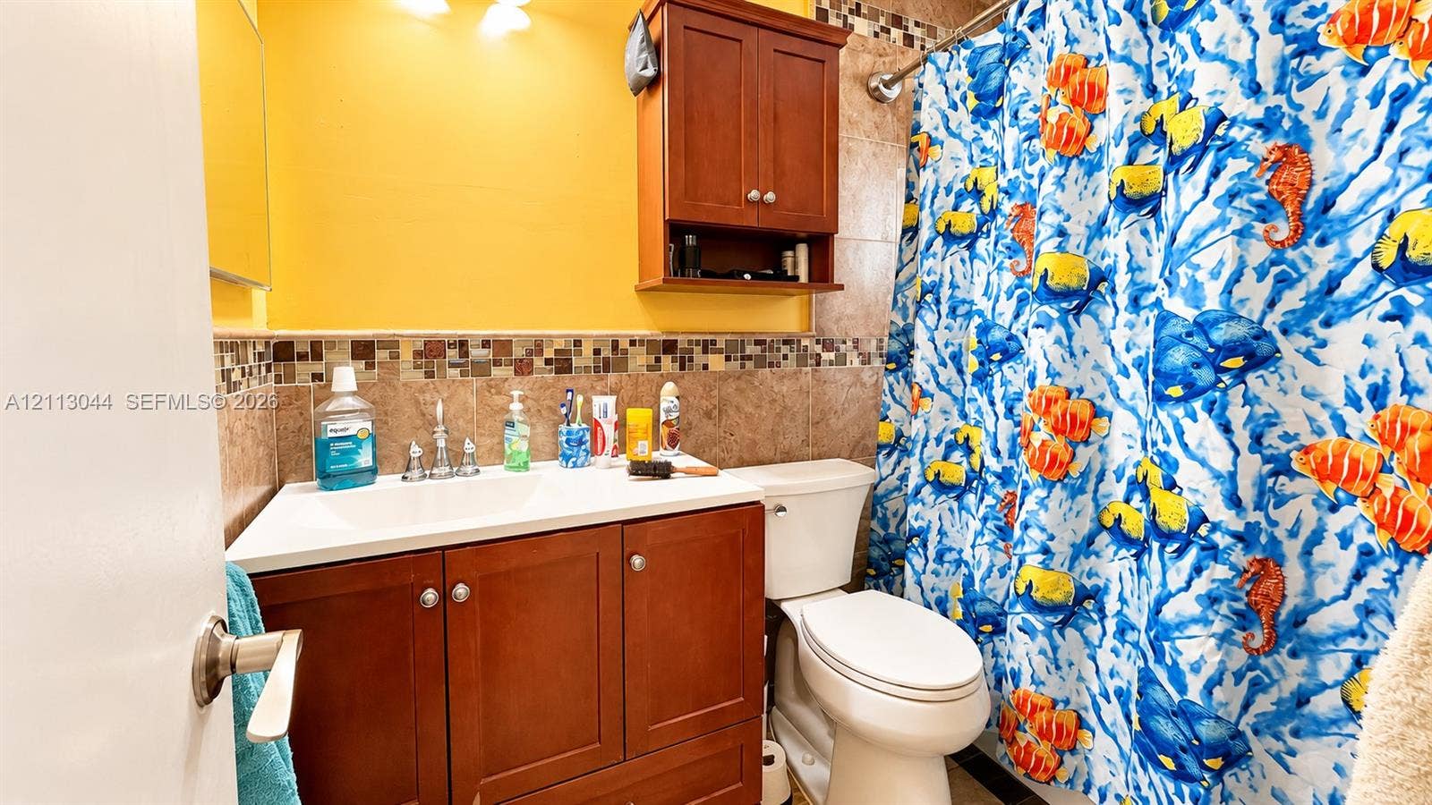 6735 NW 62nd Street, Tamarac, FL 33321 Bathroom featuring a wood-finish vanity with a white integrated sink, ceramic wall tiling with a mosaic glass accent border, and a built-in wood-finish wall cabinet